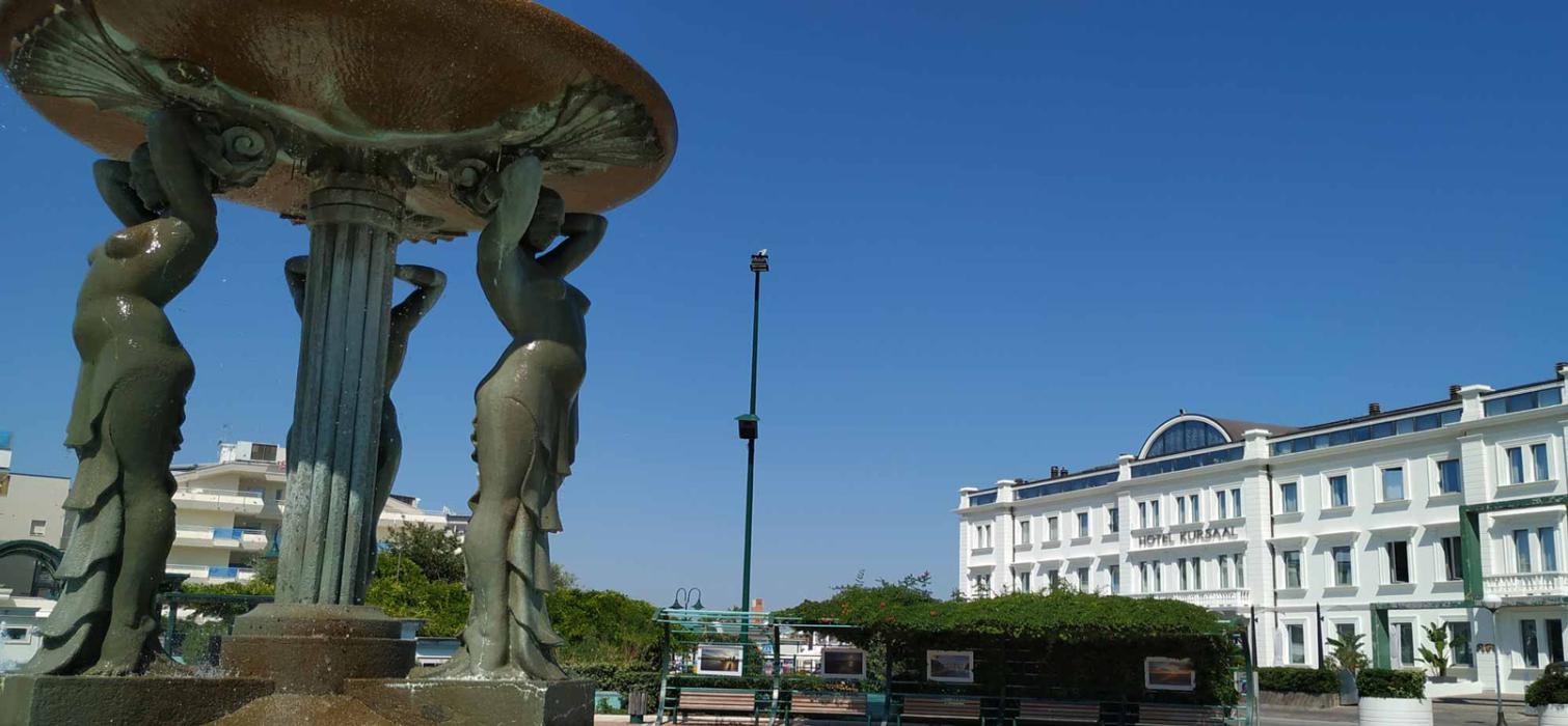 Fontana con statue vicino a un hotel bianco sotto un cielo azzurro.